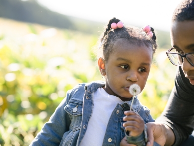 A young girl blows on a puff of dandelion seeds that she picked from the side of the road. She is with a boy, probably her older brother, enjoying the sunshine at the edge of a field. 