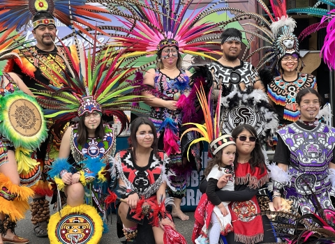 People in tradition dress are posed for a picture in a group. THe outfits are very colorful and they are wearing head-dresses with colorful feathers. 