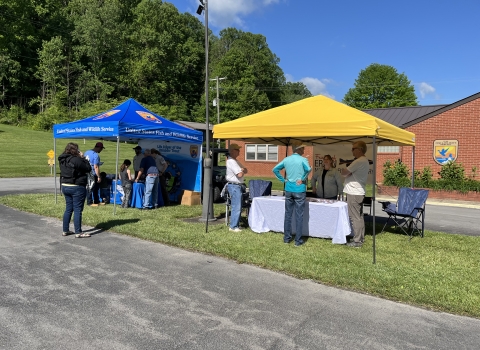 A blue USFWS canopy next to a yellow canopy. People are standing around the tables that are underneath the canopies. 