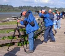 Two men in blue volunteer uniforms look intently through scopes from a boardwalk, with visitors lined up along the rail behind them.