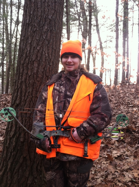 Alyssa is dress in hunting camouflaged clothes and orange vest.
