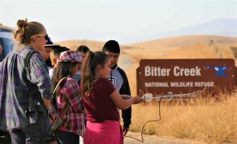 A few students hold a Yagi antenna with a female biological technician overseeing them, in front of a large brown sign on a golden grassy rolling hill, reading Bitter Creek National Wildlife Refuge.