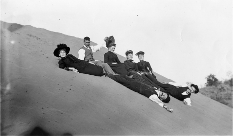 Seven people in formal clothes lounge on a sand dune in a black-and-white photo