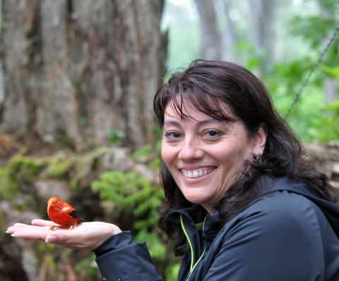 A woman in a forested setting, smiling as she holds a small red bird in the palm of her hand