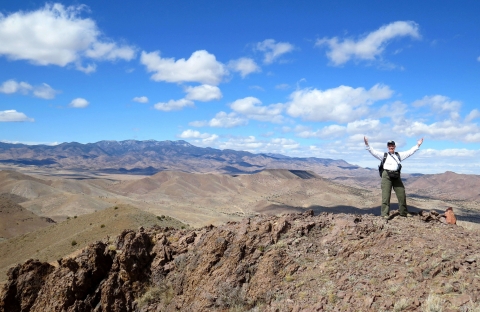 A happy person with hands raised standing on a rocky mountain peak with other mountains in the distance