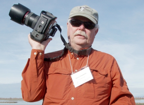 A man with gray hair and mustache wearing sunglasses and a ball cap holding his camera