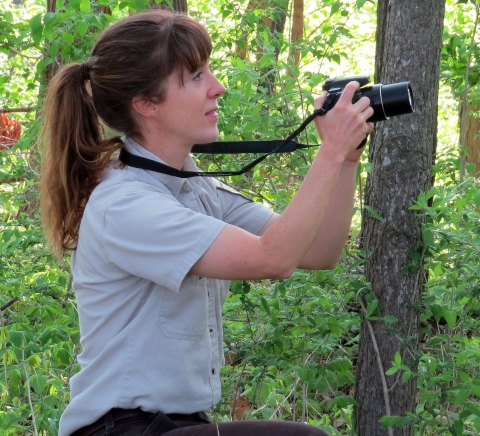 Profile shot of a woman in a forest setting crouching down and holding her camera out from her body