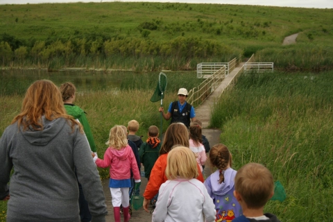 An adult leads a group of young children and moms to a bridge on the wetland in the prairie
