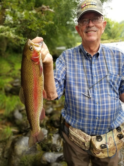 Older man in a blue plaid shirt proudly holding up a Rainbow trout for a photo