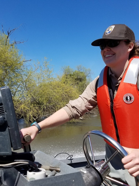 A woman drives a boat wearing a life vest and Fish and Wildlife Service hat