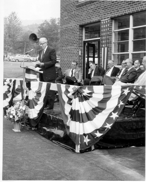 A black and white photo of a man in a suit standing before a podium in front of a brick building. A group of men sit behind him 