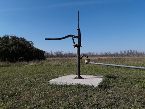 A hand pump for a well stands on a piece of concrete in a flat grassy field.