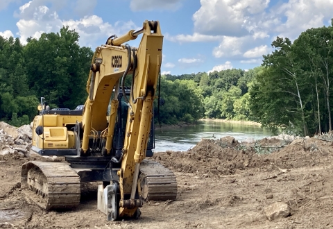 A yellow bulldozer beside the Green River on a sunny day with lots of trees along the river bank.