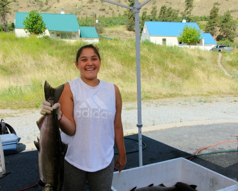 A young smiling woman in a sleeveless t-shirt holds up a salmon in her gloved right hand.