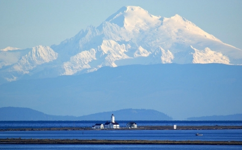 A distant view of a white lighthouse on a spit of land amid deep blue water with a snow-capped mountain looming in the background