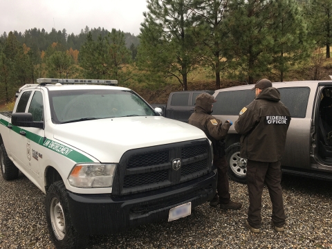 FWS Patrol Captain Kelly Knutson, a Federal Wildlife Officer, and a U.S. Forest Service agent stand next to patrol vehicles (one white truck and the other is tan) with there backs showing.