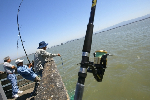 Three people fishing from a wooden pier in a photo at an odd angle with a fishing rod in the foreground