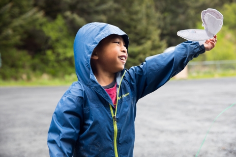 A boy in a blue raincoat holds a paper fish in the air