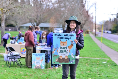 Volunteer holds a poster that reads "Winter Wildlife Field Days" with graphics of Woodland creatures. In the background are activity tables with kids activities.