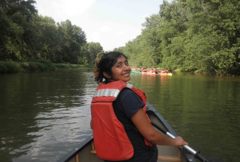 A young woman in an orange life vest paddles a canoe along a river.
