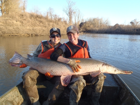 Two women hold a large alligator gar across their laps in a boat.