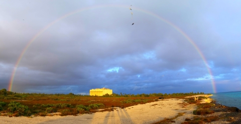 A high-arcing rainbow over a grassy beach area of an atoll in the ocean with a building in the distance