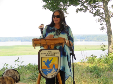 A woman with long straight grey hair wears a tie die blue shirt and sunglasses. She stands at a podium with the U.S. Fish and Wildlife Service seal. Behind her is the Rappahannock River. 
