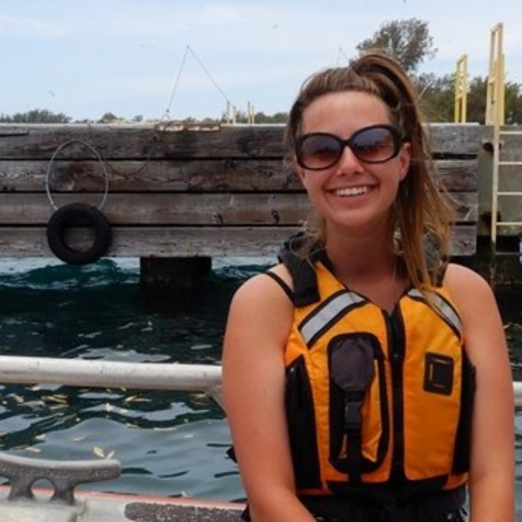 Gina McGuire looks smiles at the camera. She is wearing sunglasses and an orange lifejacket as she sits on a boat near a pier.
