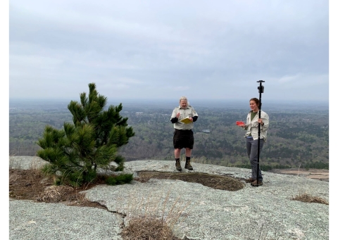 Two people stand on a sparsely vegetated granite mountain