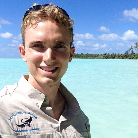 Sean Russell is wearing a Pacific Remote Islands Marine National Monument button down shirt. Behind him a crystal clear water and trees. 