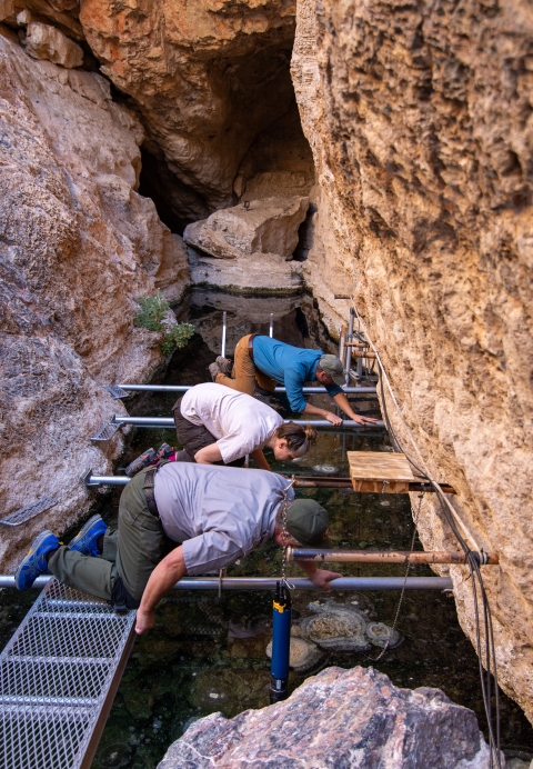 Three people kneel on a platform suspended above a water-filled crevasse and look down into the water