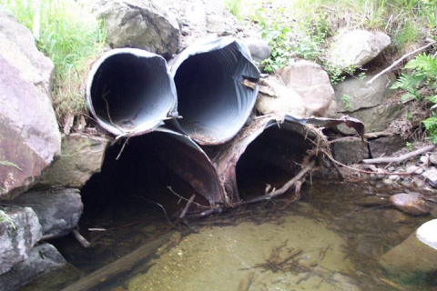 battered and rusting metal tubes (culverts) are nestled in rocks as river water covers the bottom half of the lowermost tubes