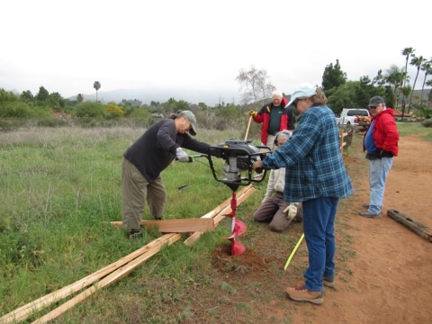 Two volunteers hold an auger from opposite sides while one refuge biologist kneels down to inspect while the auger drills into the earth. Two other volunteers stand behind the Service employee and watch. Wooden rails for fence are laid on the ground.