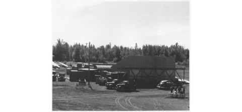 Early image of USFWS Hanger on Lake Hood Alaska with treeline in the background and vehicles parked in font. 