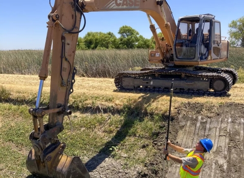 A man uses a laser level tool as a large excavator prepares to re-dig a ditch