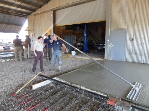 A group of people smoothing wet concrete with flat rakes across the floors in large barn.