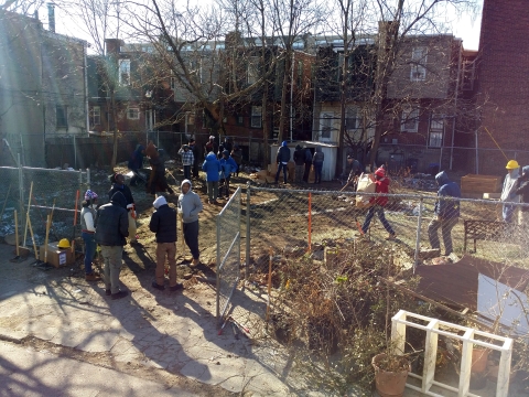 People moving debris and dead foliage out of a dirt lot with a row of houses in the background