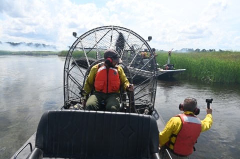 A cameraman leans off the side of an airboat to capture footage of another airboat during a prescribed fire