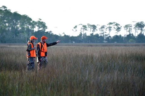 A man and a woman dressed in bright orange safety gear walk across a saltmarsh during a deer hunt