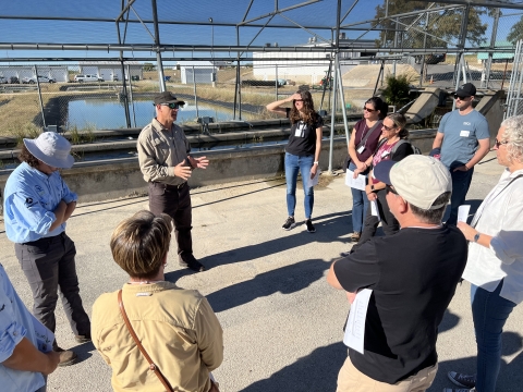 This image shows a man in a USFWS uniform in front of a pond and fish raceway, talking to a group of nine visitors outside in the morning sun on a bright day.