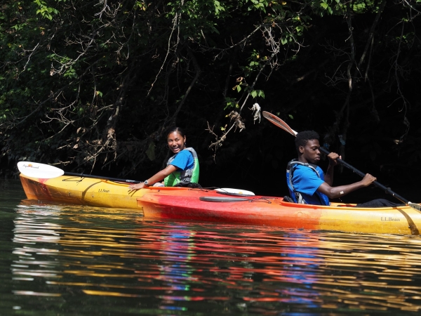 Two young people are smiling and paddling on bright kayaks. 