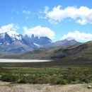 Mountain range with snow-capped peaks in background, and body of water, grass-covered hills, and field in foreground