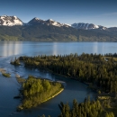 An aerial landscape photo of a large blue lake along forested land, with snow-capped mountains in the background.