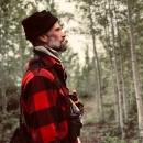 A side profile from the waist up, of a person wearing a red flannel shirt, a bear spray holster around their torso, and a black knit hat. They look away from the camera and off-frame with trees behind them.. 