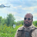 A man in sunglasses wearing a U.S. Fish and Wildlife Service uniform standing in a green field with a helicopter overhead