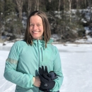 A woman with shoulder-length brown hair wearing a blue jacket and gloves smiles in front of a forested hill with snow on the ground.