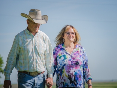 A man wearing a cowboy hat and a woman walk hand-in-hand across a grassy field