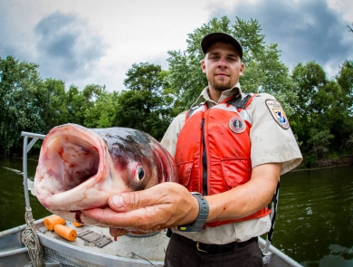 A person in a Service uniform and life vest holds a large hideous fish with bulging eyes and a gaping mouth up the camera. 