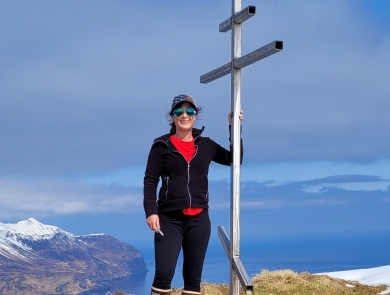 A woman wearing a hat and sunglasses on a high summit overlooking snowcapped mountains and ocean. She stands next to a metal structure.