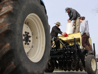 Three people standing on a tractor-pulled seed planter, with a large tractor tire in the foreground.